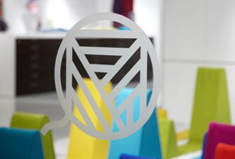 Gravürlü cam tipi çizgili logolu şeffaf cam pencere, arkada renkli şekiller var.
